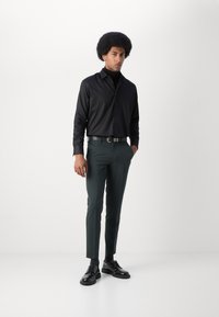 Camisa preta de manga longa, calças justas verde-escuras, cinto preto e sapatos pretos brilhantes. Design minimalista com uma textura suave.
