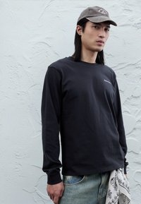 Camiseta negra de manga larga con cuello redondo y un pequeño logo blanco. Combinada con jeans azul claro y un pañuelo blanco con estampado en la cintura.