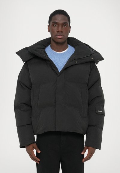 Chaqueta acolchada negra con un alto cuello, forma rectangular y costuras horizontales. Presenta bolsillos laterales y un parche con el logo en la manga.