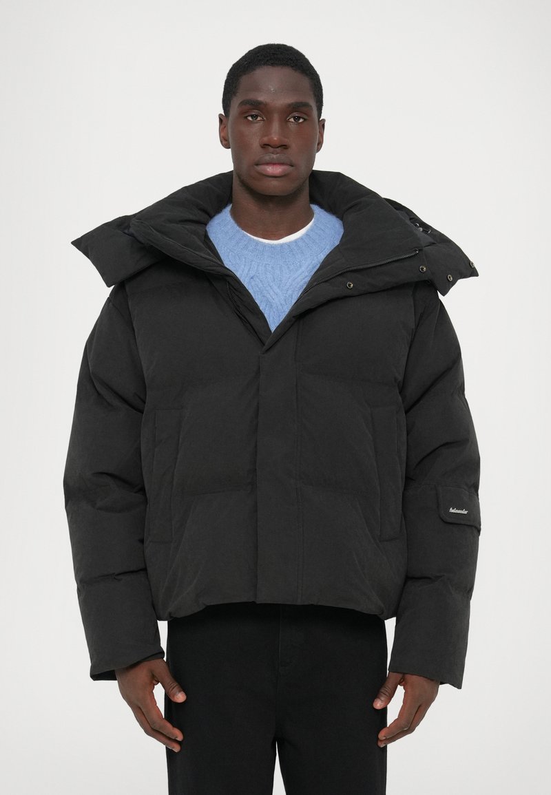 Black padded jacket with a high collar, boxy shape, and horizontal stitching. Features side pockets and a logo patch on the sleeve.