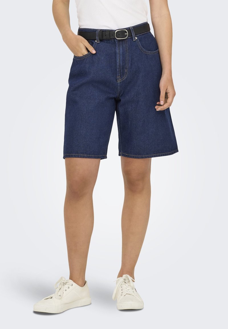 Shorts en denim à coupe décontractée, de couleur bleu foncé, avec poches avant et une ceinture noire. Associés à des baskets blanches et un haut blanc ajusté.