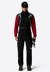 Juodo ir raudono spalvų sportinis aprangos rinkinys, kuriame yra berankovė striukė, aptemptos kelnės ir kepurė, papildyti diržu ir žygio lazdomis.