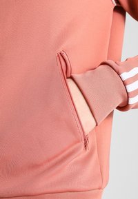 Pink athletic sweatshirt featuring a zippered pocket; fabric appears smooth with ribbed cuffs and three white stripes on the sleeve.