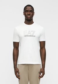 Λευκό βαμβακερό T-shirt με κοντά μανίκια, με μαύρο κεντημένο λογότυπο "EA7 EMPORIO ARMANI" στο κέντρο του στήθους. Λείος υφασμάτινος τόνος.
