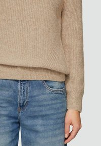 Beige Strickpullover mit strukturiertem Muster, leicht oversized geschnitten und mit gerippten Bündchen, kombiniert mit mittelblauen Jeans aus Denim mit klassischer Tasche.