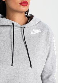 Šedá mikina s kapucí a stahovací šňůrkou, s bílým logem Nike na hrudi a textem podél rukávů. Měkká tkanina s volným střihem.
