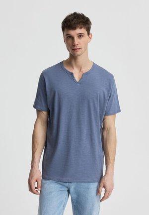 Giovane uomo con una maglietta henley blu a maniche corte e jeans azzurri, in piedi davanti a uno sfondo chiaro e semplice.