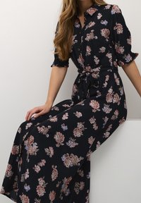 Maxi mekko, jossa on tummansinisestä kankaasta valmistettu kukkakuosi, jossa on vaaleanpunaisia ja sinisiä sävyjä. Napitettavat etuosat ja solmittava vyötäröyksityiskohta.