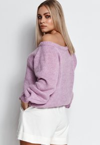 Fioletowy sweter dzianinowy z szerokim dekoltem typu off-the-shoulder oraz obszerne rękawy, w zestawieniu z białymi szortami, prezentujący luźny krój i strukturę.
