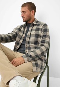 Beige og sort ternet flannel skjorte med knaplukning og brystlommer; båret over en mørkegrå sweater og parret med corduroybukser.