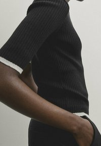 Top corto a maniche corte nero a coste con dettagli a contrasto in bianco sui polsini, design aderente, texture morbida e silhouette leggermente cropped.