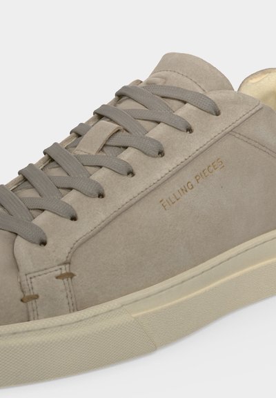 Gros plan d’une basket en daim beige clair avec lacets assortis et semelle en caoutchouc crème, portant le texte "Filling Pieces" sur le côté.