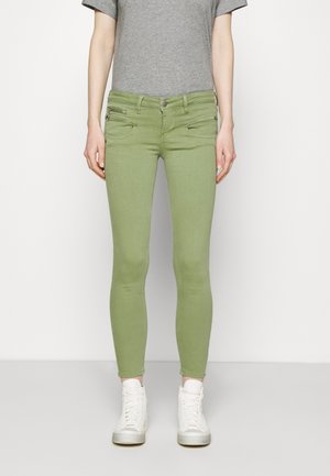Green skinny jeans made of denim, featuring two front pockets with zippers, paired with a gray T-shirt and white high-top sneakers.