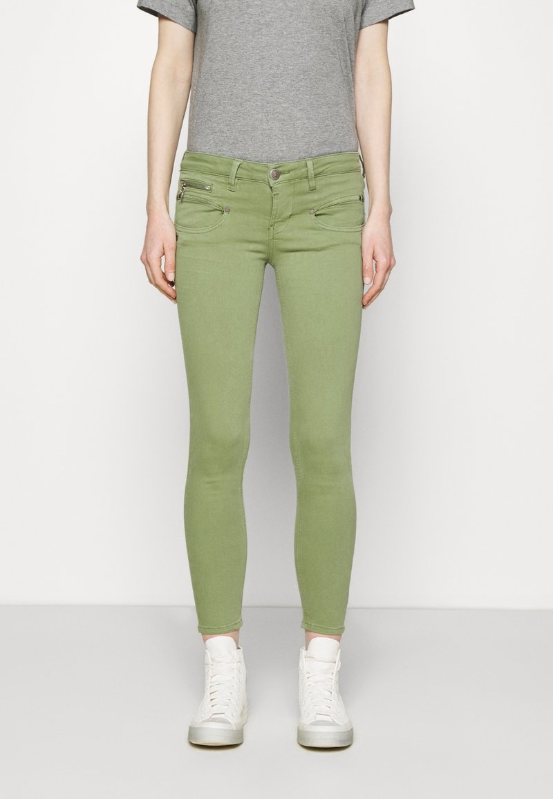 Green skinny jeans made of denim, featuring two front pockets with zippers, paired with a gray T-shirt and white high-top sneakers.