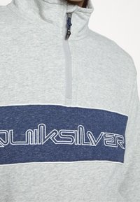 Sudadera de media cremallera en gris claro con una franjas horizontal en azul marino a través del pecho y texto en blanco contorneado que dice "Quiksilver."