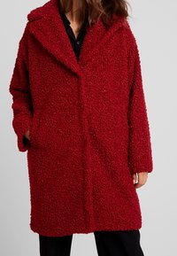 Červený, texturovaný, chlupatý kabát s výřezovým límcem, volným oversized střihem a předními kapsami. Má dlouhé rukávy a měkký, útulný vzhled.