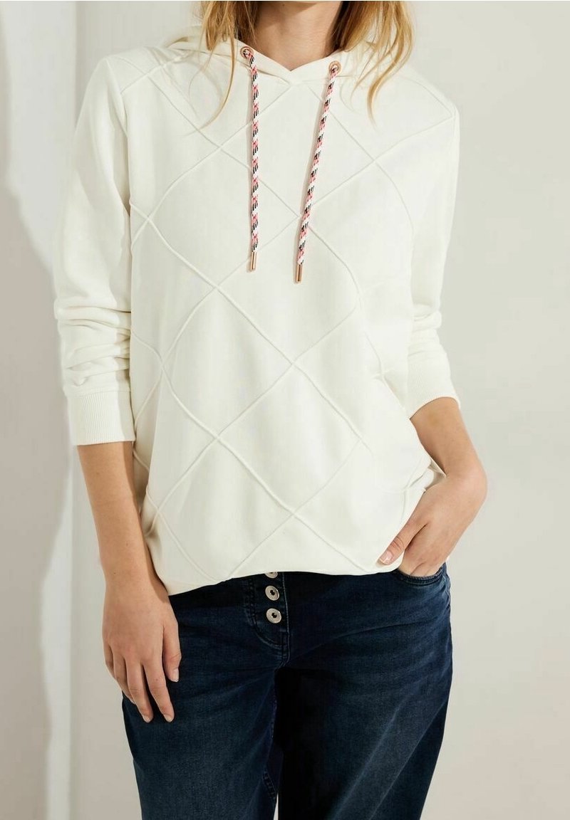 Femme portant un sweat à capuche matelassé blanc avec des cordons à motifs et un jean bleu foncé avec trois boutons visibles, main dans la poche.