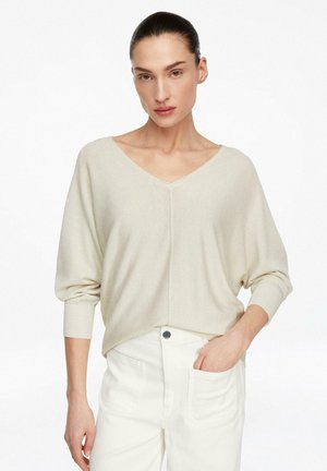 Kobieta o ciemnych włosach w kremowym swetrze z dekoltem w serek, włożonym w białe spodnie z wysokim stanem, jedna ręka w kieszeni, neutralne tło.