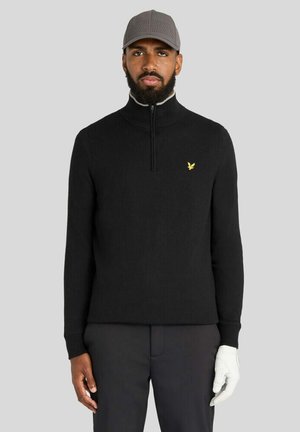 Mand iført sort lynlås-sweater med gult logo, mørke bukser, grå kasket og hvid golfhandske på højre hånd mod ensfarvet baggrund.