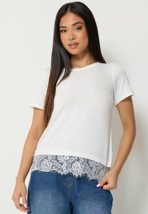 Joven mujer con pelo largo y oscuro que lleva una camiseta blanca de manga corta con encaje y vaqueros azules, de pie frente a un fondo liso.