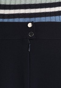 Vêtement noir avec une ceinture en tricot rayée bleue, grise et noire. Équipé d'une fermeture éclair et de boutons-pression métalliques.
