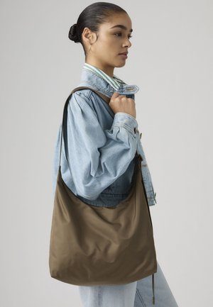 Νεαρή γυναίκα με σκούρα μαλλιά σε κότσο, φορώντας τζιν μπουφάν και κρατώντας μεγάλη καφέ τσάντα ώμου, σε απλή φόντο.