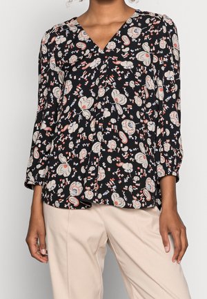 Femme portant un chemisier noir avec des motifs floraux et géométriques abstraits en blanc, rouge et bleu, associé à un pantalon beige clair.