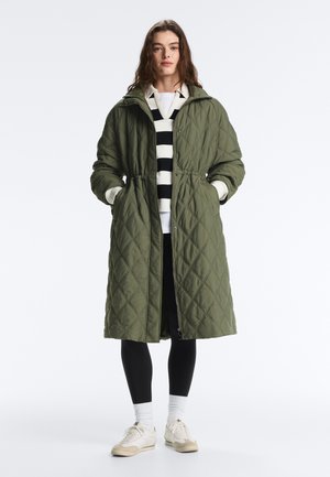 Frau trägt einen langen olivgrünen gesteppte Mantel über einem schwarz-weiß gestreiften Shirt, schwarzen Leggings, weißen Socken und beigen Turnschuhen.