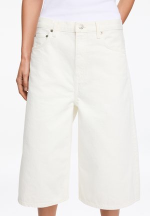 Kvinde iført hvide denimshorts med brede ben til knæet, med knapper foran og lommer, kombineret med en hvid top.