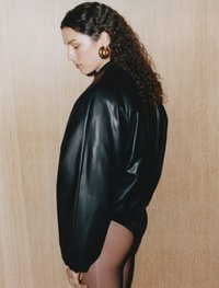 Zwarte leren oversized jas met een gladde textuur, gedragen over donkere shorts. Model heeft krullend haar en grote gouden oorbellen.