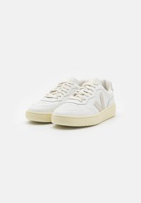White leather sneakers with a textured surface, flat laces, light beige accents, and a soft, chunky sole. Classic sneaker silhouette.