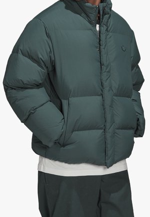 Person iført en mørkegrøn polstret jakke med hænderne i lommerne, over en hvid skjorte og matchende mørkegrønne bukser.