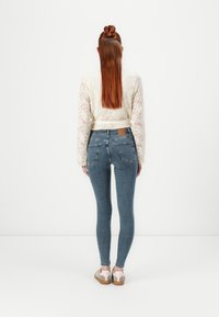 Jeans ajustados en denim azul medio, con bolsillos traseros y una etiqueta de cuero marrón. Combinados con una blusa de encaje crema, que presenta mangas largas y una cintura ceñida.