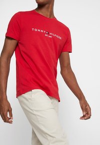 Camiseta de algodón roja con mangas cortas, que presenta un logo blanco y el texto "TOMMY HILFIGER EST. 1985" centrado en el pecho. Ajuste de longitud media.