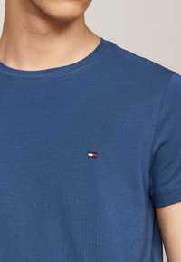 Camiseta azul de suave tejido de algodón con cuello redondo. Presenta un pequeño logotipo bordado en rojo, blanco y azul en el lado izquierdo del pecho.