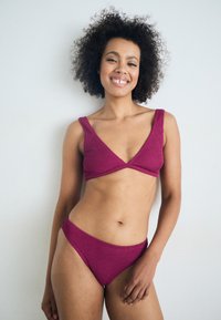Magenta juostelėmis bikinis rinkinys, kuriame yra trikampio formos liemenėlė su plačiomis petnešėlėmis ir žemai nuleistais apatiniais. Lygus audinys ir minimalus dizainas.