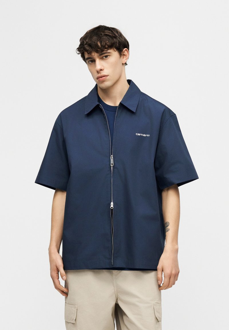 Jeune homme portant une chemise zippée à manches courtes bleu marine et un pantalon cargo beige, debout avec une expression neutre sur un fond uni.