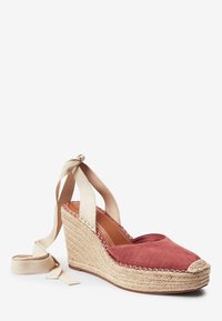 Espadrille compensée avec un dessus en suède rouge, des brides en tissu beige à la cheville, et une semelle plateforme en jute. Design accrocheur avec un bout en cap.
