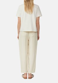 Hell gestreiftes Kurzarmoberteil in Creme, kombiniert mit locker sitzenden beigen Hosen. Die Schuhe sind flache Sandalen mit einer Schleifenverzierung.