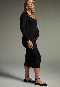 Černé žebrované šaty s dlouhým rukávem, přiléhavý střih, délka ke kolenům, nošeny s černými balerínami s bílými puntíky a kotníkovými řemínky.