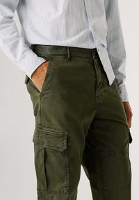 Groene cargobroek van zacht materiaal, met zijzakken en een ontspannen pasvorm, gedragen met een lichtblauw gestreept overhemd.