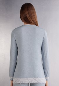 Lichtblauw langslijvende top van zacht materiaal met een ronde halslijn en decoratieve witte kantafwerking aan de onderrand en de manchetten.
