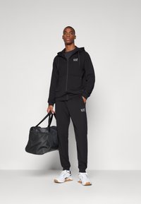 Black zip-up hoodie and matching joggers with EA7 logo. Model holds a black duffel bag. White sneakers with gum sole complete the look.