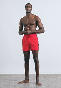 Piros fürdőnadrág, könnyű anyagból, rugalmas derékpánttal és húzózsinórral. Az oldalon kontrasztos fekete-fehér márkajelzés látható.