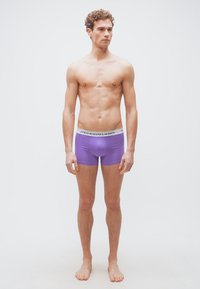 Νεαρός άνδρας με σγουρά μαλλιά, όρθιος ξυπόλητος, φοράει μωβ boxer briefs Polo Ralph Lauren μπροστά σε απλό φωτεινό φόντο.