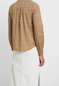 Chemise à manches longues à motifs marron avec un détail froncé dans le dos, portée sur une jupe blanche avec une fente latérale et des coutures visibles.