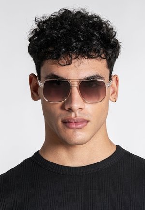 Ung mand med krøllet sort hår iført store tonede solbriller og en sort ribt shirt mod en ensfarvet lys baggrund.