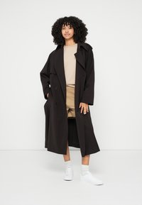 Dolgi črn plašč s prekrivanjem na dvojni gumb, stranskimi žepi in širokim ovratnikom, nošen čez svetel pulover in bež kratke hlače.