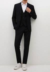 Completo nero su misura con rever a scollo a punta, camicia bianca e gilet a tre bottoni. Abbinato a scarpe da tennis bianche per un tocco informale.