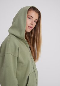 Felpa con zip verde oliva realizzata in materiale morbido, dotata di un ampio cappuccio, tasche frontali e dettagli di cucitura lungo le cuciture.